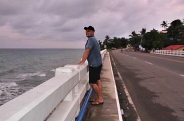 A man standing on a bridge against a gray sky and raging sea and looking into the distance