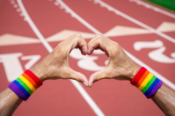 Gay athlete wearing rainbow pride wristbands making love heart hands gesture against a red athletic track background - 443158430