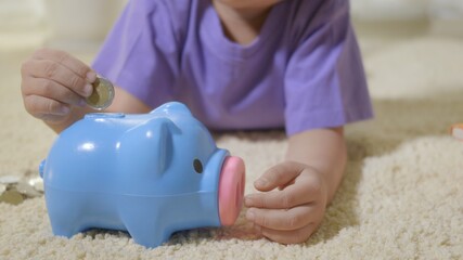 Happy Asian kid boy preschool putting pin money coins into blue faced piglet slot. Little child putting coin into piggy bank for saving with pile of coins at home, Investment education
