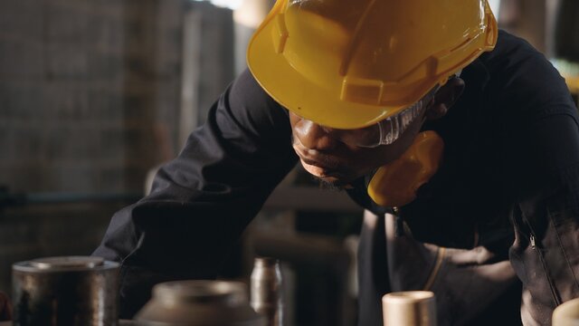 American Industrial Black Young Worker Man With Yellow Helmet And Ear Protection Surveillance For Examination In Front Machine, Engineer Inspection Checking Work In The Industry Factory.