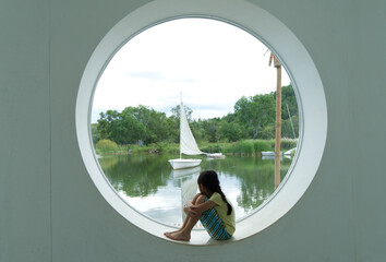 Asian girl sits looking at the view of a sailing boat from a circular aperture
