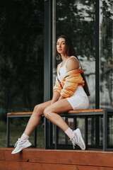 Fototapeta premium Fashion portrait of young trendy woman dressed in white dress, jacket and sneakers sitting on the bench against city cafe, summer street fashion. portrait of joyful woman