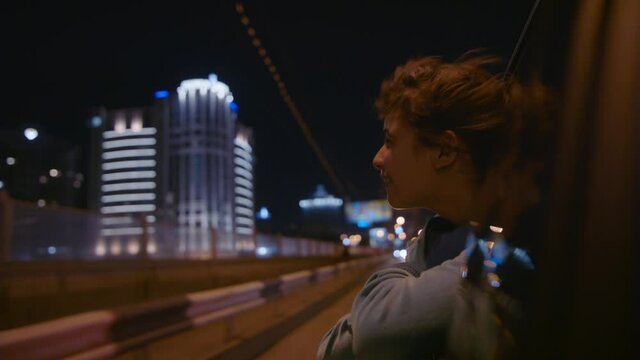 Kid Tourist Traveler Looks Out Of Car Window At Lights Of Night City. Little Girl Enjoying Journey. Child Leaned Out Of Car Window While Driving And Enjoys Trip Around City At Night, Laughs And Smiles