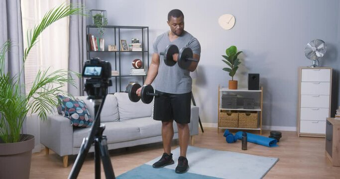 Bodybuilder. Fitness muscled multiracial man standing and lifting dumbbell at his apartments. Beautiful sporty guy male power concept. Stock video