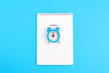 Classic alarm clock and empty notepad on blue background. Top view, copy space.