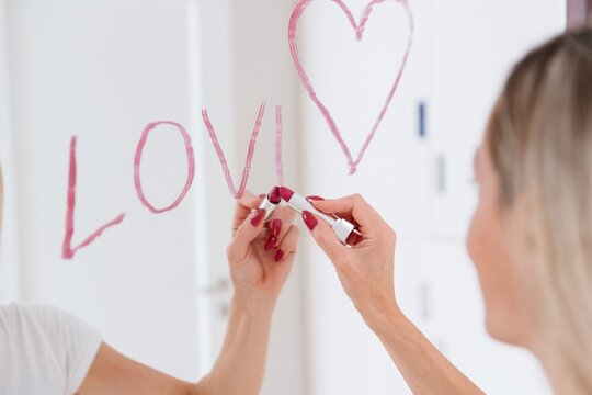 Blonde woman drawing heart in lipstick on mirror.