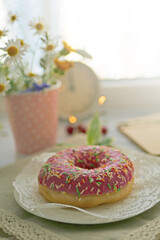 Donut in pink glaze on a background of wild flowers of chamomiles, fresh berries on a wooden table