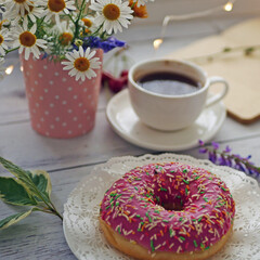 Summer breakfast with a cup of coffee and a donut in pink glaze on a background of wild flowers of chamomiles, fresh berries, an alarm clock on a wooden table.Home cozy summer rustic background