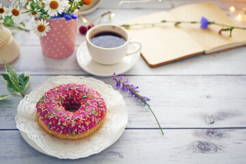 Summer breakfast with a cup of coffee and a donut in pink glaze on a background of wild flowers of chamomiles, fresh berries, an alarm clock on a wooden table.Home cozy summer rustic background