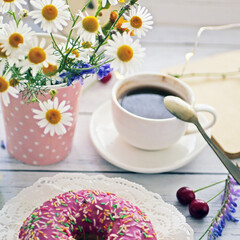 Summer breakfast with a cup of coffee and a donut in pink glaze on a background of wild flowers of chamomiles, fresh berries, an alarm clock on a wooden table.Home cozy summer rustic background
