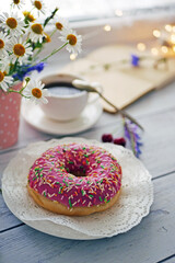 Summer breakfast with a cup of coffee and a donut in pink glaze on a background of wild flowers of chamomiles, fresh berries, an alarm clock on a wooden table.Home cozy summer rustic background