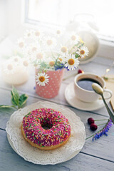 Summer breakfast with a cup of coffee and a donut in pink glaze on a background of wild flowers of chamomiles, fresh berries, an alarm clock on a wooden table.Home cozy summer rustic background