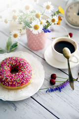 Summer breakfast with a cup of coffee and a donut in pink glaze on a background of wild flowers of chamomiles, fresh berries, an alarm clock on a wooden table.Home cozy summer rustic background