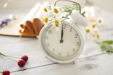 Summer time background. White alarm clock arrows noon on a wooden table around wildflowers fresh berries. Summer background concept, copy space