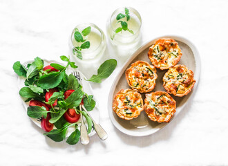 Aperitif table - mini quiche with salmon and zucchini, fresh tomato arugula salad and mint cocktail on a light background, top view