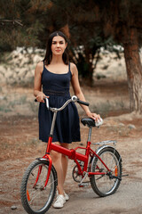 Naklejka premium a young brunette woman of Asian appearance in a dark blue dress is standing next to a bicycle with a punctured wheel. there is a grain and a small depth of focus in the photo for artistic purposes