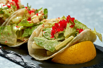 Mexican food dish - tacos with salmon, lettuce, white sauce and tobiko caviar on a black plate on a blue background