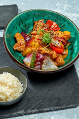 Appetizing pan-Asian dish - chicken wok in sweet and sour sauce with bell pepper, onion, sesame seeds and rice garnish in a blue bowl on a gray background