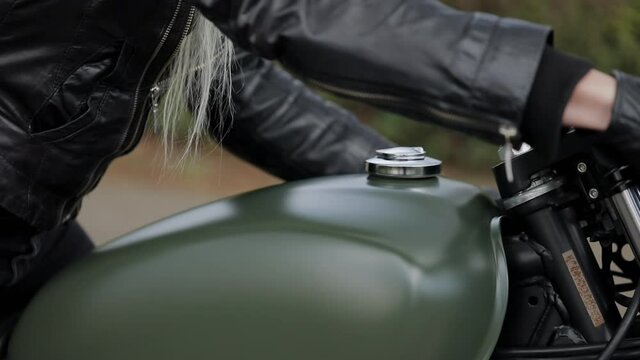 Blonde lady biker in black pants and leather jacket sits on large motorbike and takes handlebars before race extreme closeup