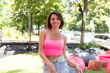 Beautiful woman in bright pink top and casual jean shorts wears beaded necklace in summer outdoor cafe positive smile enjoying summer cheerful