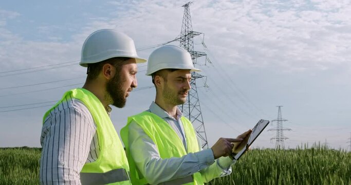 Employees in green vests and hardhats with tablet test power transmission lines standing in field on spring day slow motion
