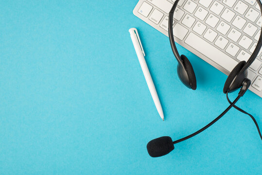 Top view photo of wired black headset pen and white keyboard on isolated pastel blue background with blank space