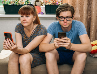 Photo of modern family relationships. Mom and teenage son look at their phones at home while sitting on the sofa - 443200453
