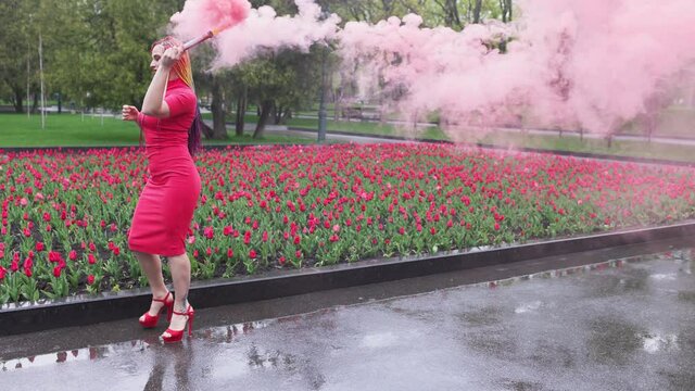 A girl with makeup with rainbow braids in red dress posing in red smoke against the background of a blooming flower bed