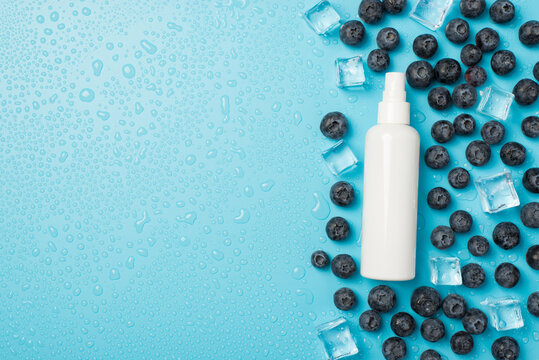 Above photo of blueberries cubes ice water drops and white bottle of spray isolated on the blue background with copyspace