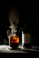 Great view of old-fashioned glass of cocktail and bottle in dark with white smoke around
