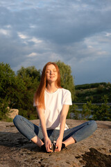 Nature, wellbeing, Relaxation, meditation and mental health. Interacting, Thriving with nature. Calming and enchanting effect. Young redhead woman sitting on rock and watching on river on sunset