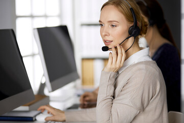 Friendly businesswoman talking by headset while sitting in office. Call center and diverse people group in business