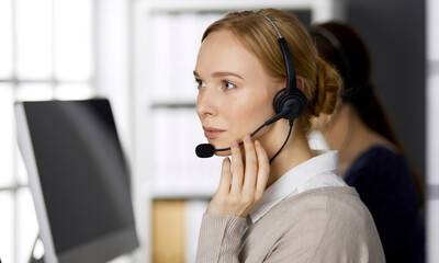 Friendly businesswoman talking by headset while sitting in office. Call center and diverse people group in business