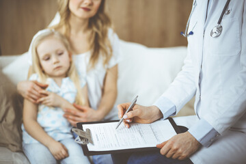Doctor and patient. Pediatrician using clipboard while examining little girl with her mother at home. Sick and unhappy child at medical exam. Medicine concept