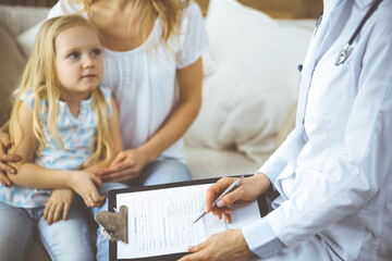 Doctor and patient. Pediatrician using clipboard while examining little girl with her mother in clinic at home. Happy cute caucasian child at medical exam. Medicine concept