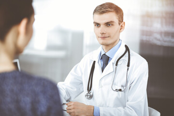 Doctor man, with a stethoscope, is listening to his patient, while sitting at the desk in the sunny cabinet at hospital. Perfect medical service in clinic. Happy future in medicine and healthcare