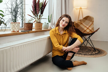Cute young woman relaxing on the floor near a warm radiator - obrazy, fototapety, plakaty