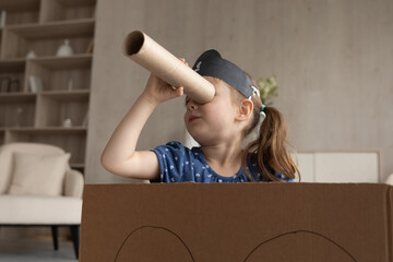 Cute adorable little girl in black pirate hat looking in spyglass, sitting in toy cardboard ship,...