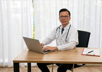 Fototapeta premium Asian young male happiness doctor typing laptop sitting at desk in hospital room