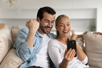 Excited young couple looking at phone screen together, celebrating success close up, sitting on...