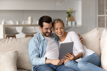 Happy young couple using computer tablet, hugging relaxing on couch at home together, smiling woman and man in glasses looking at modern device screen, browsing apps, shopping or chatting online