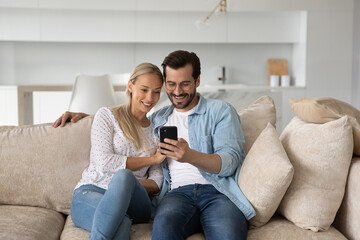 Happy young couple man and woman using smartphone together, hugging, sitting on cozy couch at home,...