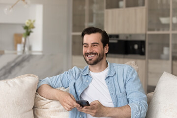 Overjoyed laughing young man distracted from smartphone sitting on couch, holding phone in hands,...