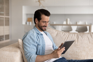 Fototapeta premium Smiling man in glasses using tablet, sitting on cozy couch at home, positive happy young male looking at modern device screen, typing writing message in social network having fun leisure time