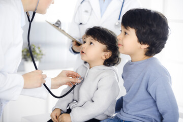 Woman-doctor examining a child patient by stethoscope. Cute arab toddler and his brother at physician appointment. Medicine concept