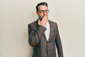 Middle age man with beard and grey hair wearing business jacket and glasses bored yawning tired covering mouth with hand. restless and sleepiness.