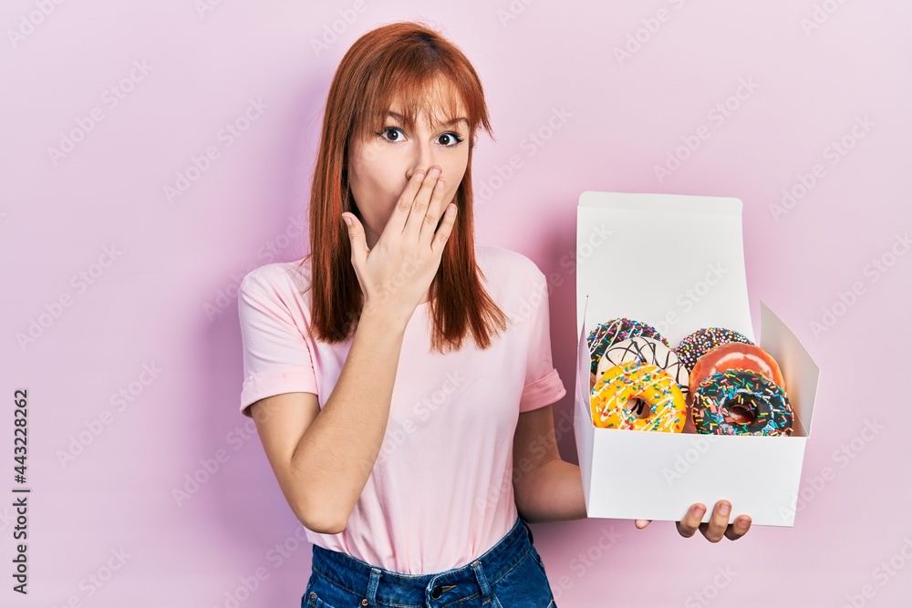 Poster Redhead young woman holding tasty colorful doughnuts covering mouth with hand, shocked and afraid for mistake. surprised expression