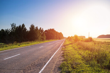 Fototapeta premium Asphalt road among green grass on summer sunset.