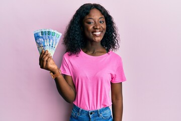 Beautiful african young woman holding south african 100 rand banknotes looking positive and happy standing and smiling with a confident smile showing teeth