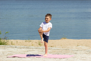 Fototapeta na wymiar A little boy in a white t-shirt and blue shorts does a warm-up on the background of the sea. Child does gymnastic exercises. Healthy lifestyle. Warm sunny summer day. Sport and activity. Stretching